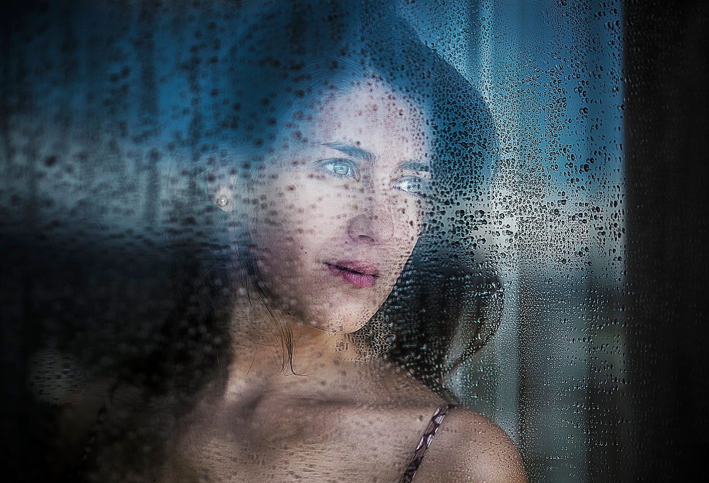 Dark haired woman looking through a rainy window with trepidation