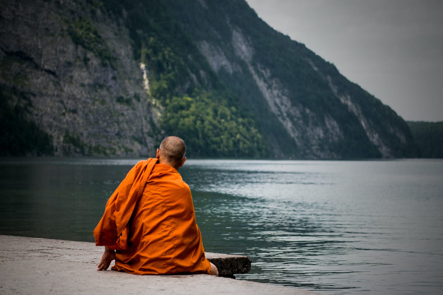 A monk, sitting by a lake. He looks over to the mountainside.