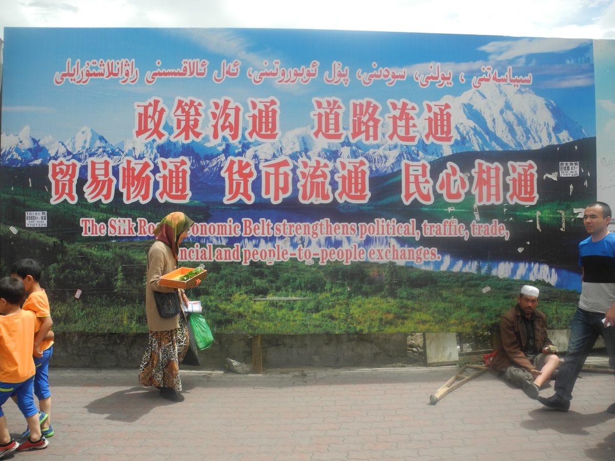 A peddler and beggar in front of a propaganda poster for the Silk Road Economic Belt in Urumqi, in China's far western Xinjiang region. Image: Wade Shepard.