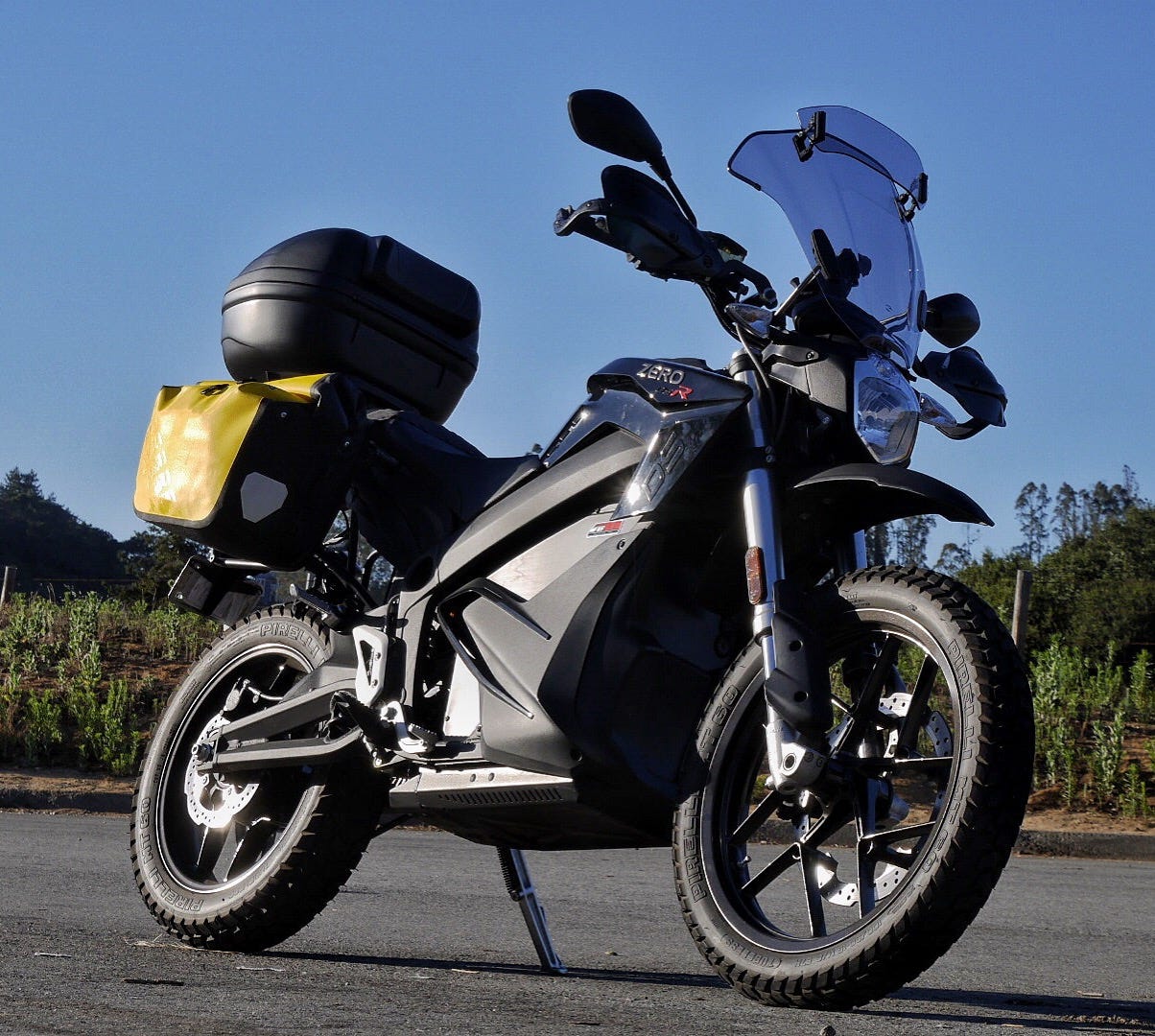 A black, electric motorcycle, parked on asphalt in the late afternoon. The motorcycle is a Zero DSR, an adventure style motorcycle, with a large 19" front tire with aggressive tread, and a 17" rear tire with similar tread. The motorcycle has a single headlight, hand protectors, and a large, two-piece windshield. A large black box hangs from the frame where a gas-powered engine usually sits. That is the battery. Two yellow and black saddlebags sit on the back of the bike, along with a hard-shelled topcase on the back. The bike is modern and contemporary in style
