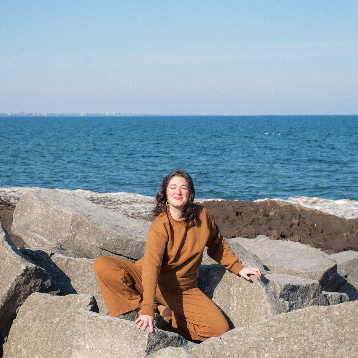 phoebe sitting in the rocks along the bech. She is crouching down in a brown sweat suit smiling and squinting in the sun, her cheeks are rosy from the wind and cold