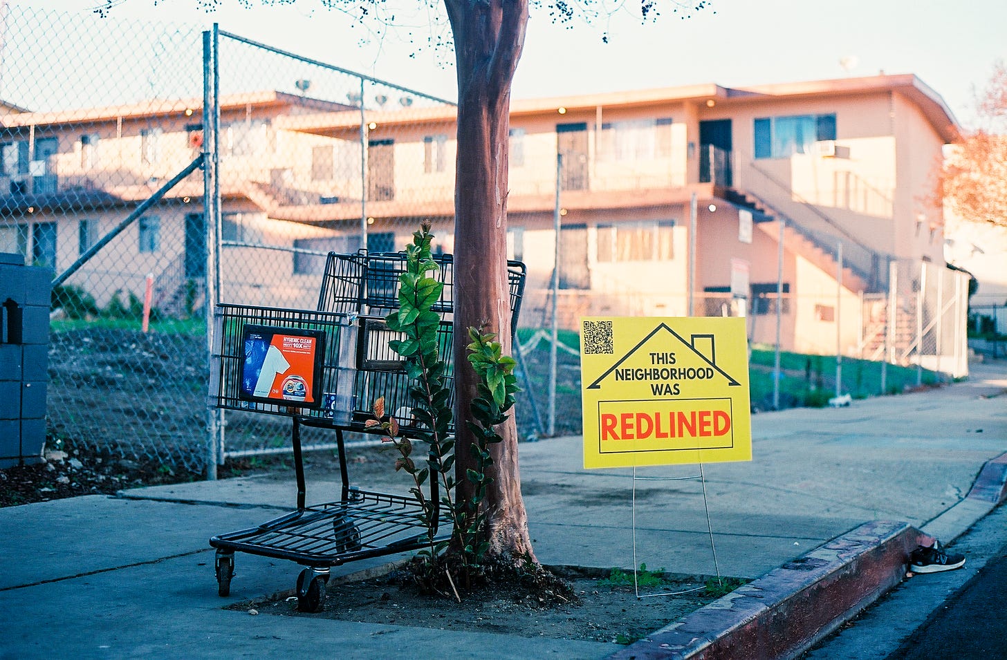 Photograph of an empty parking cart on a sidewalk in front of a chainlink fenced off empty lot next to a tree with one of my yard signs under the tree.