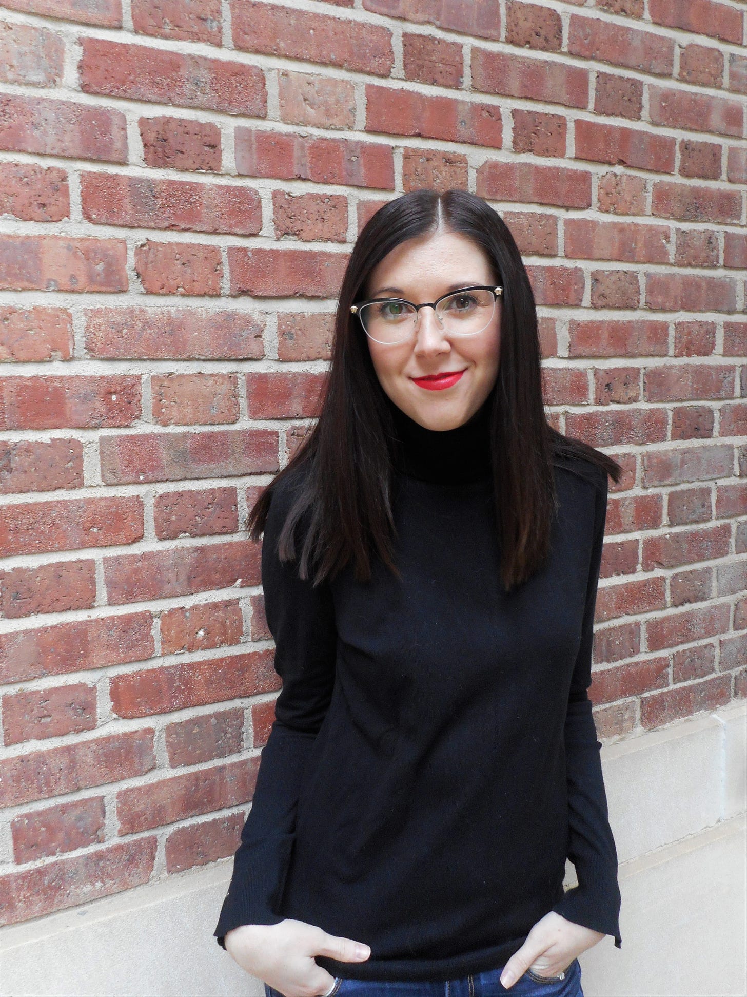 Photo of author Sarah Fawn Montgomery from the hips up, standing in front of a brick wall and wearing a long-sleeve black shirt and glasses.