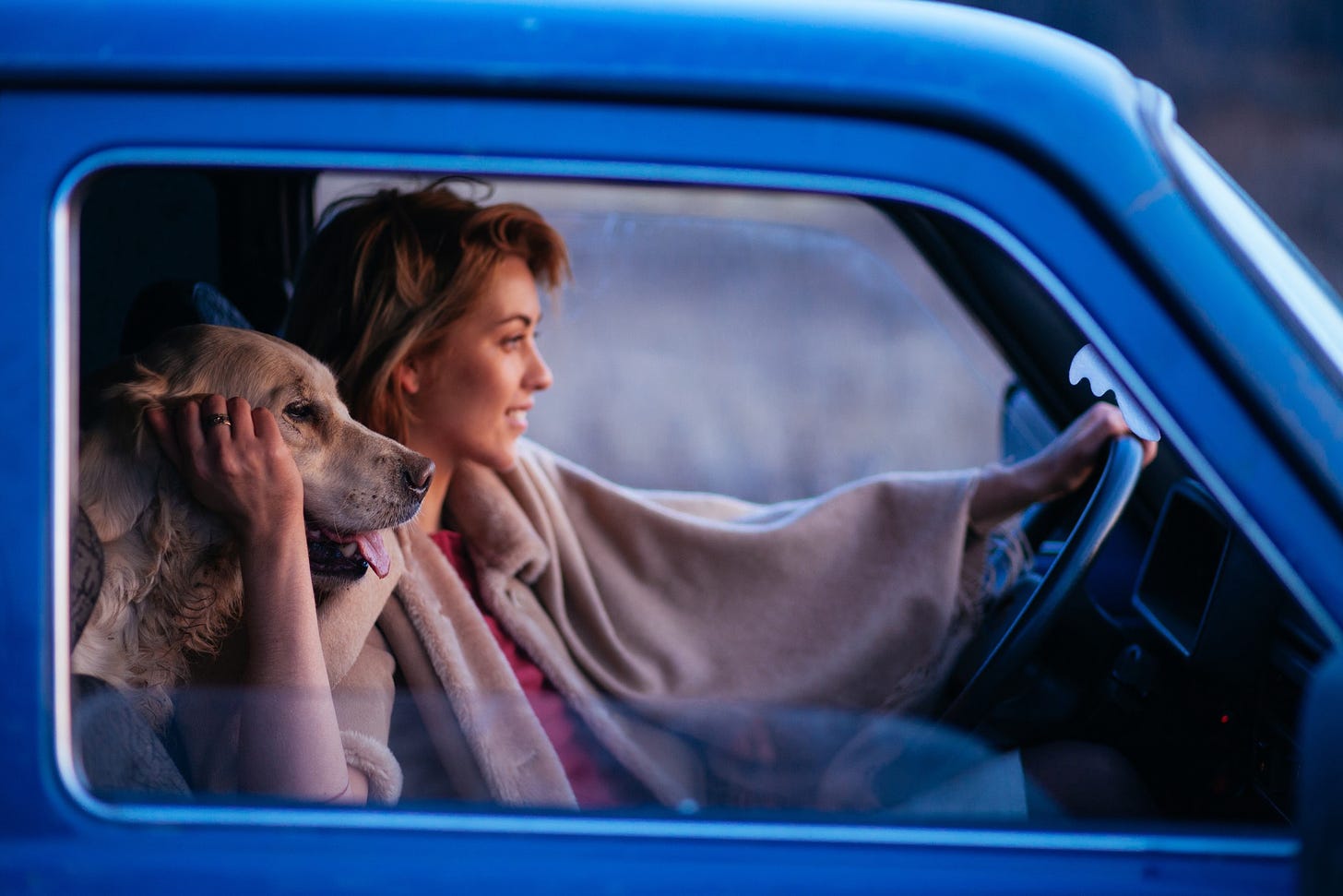 A woman in a blue truck smiles as she holds her steering wheel and pets her dog, whose head rests on her shoulder.