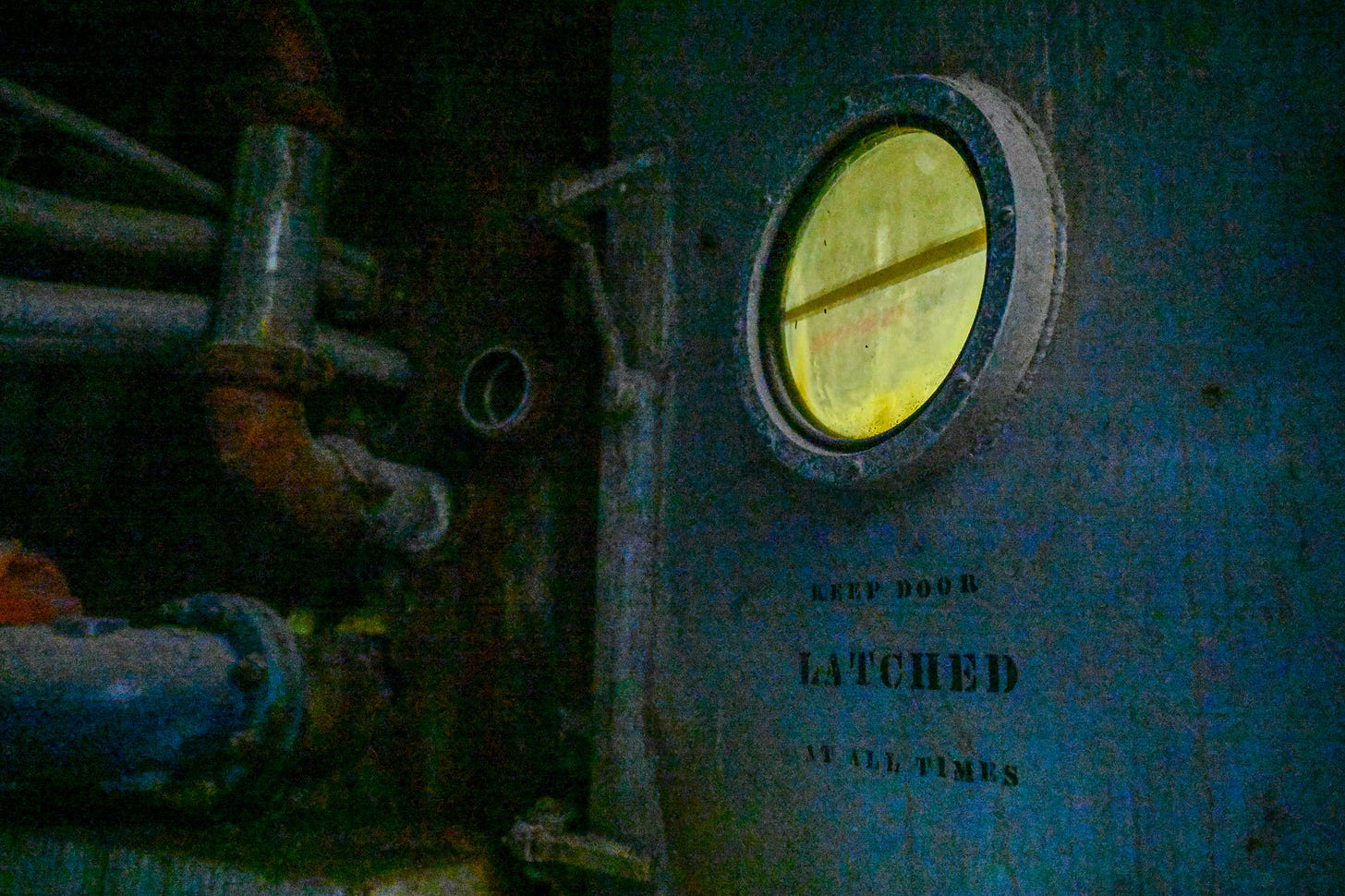 a dim image of a large steel door. the light source comes from the other side of the door visible through a round porthole. stenciled on the door are the words in all caps "keep door latched at all times"