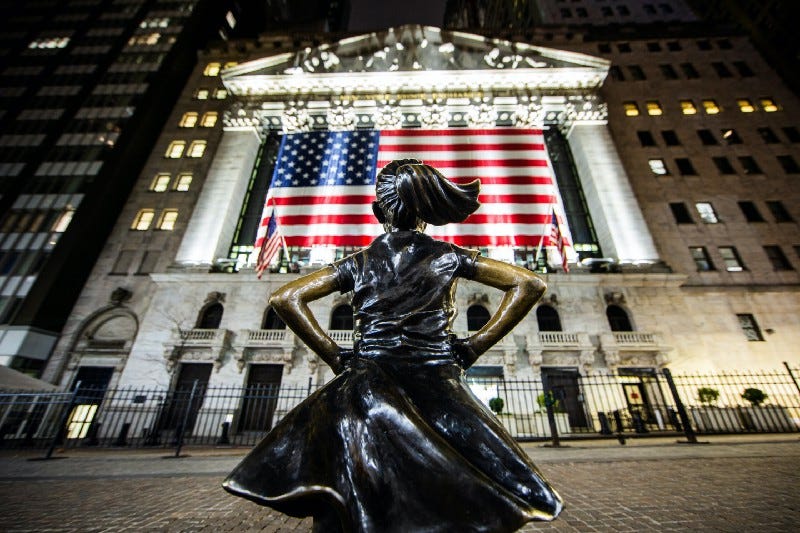 A statue of a girl with her hands on her hips, facing an American flag draping from the New York Stock Exchange.