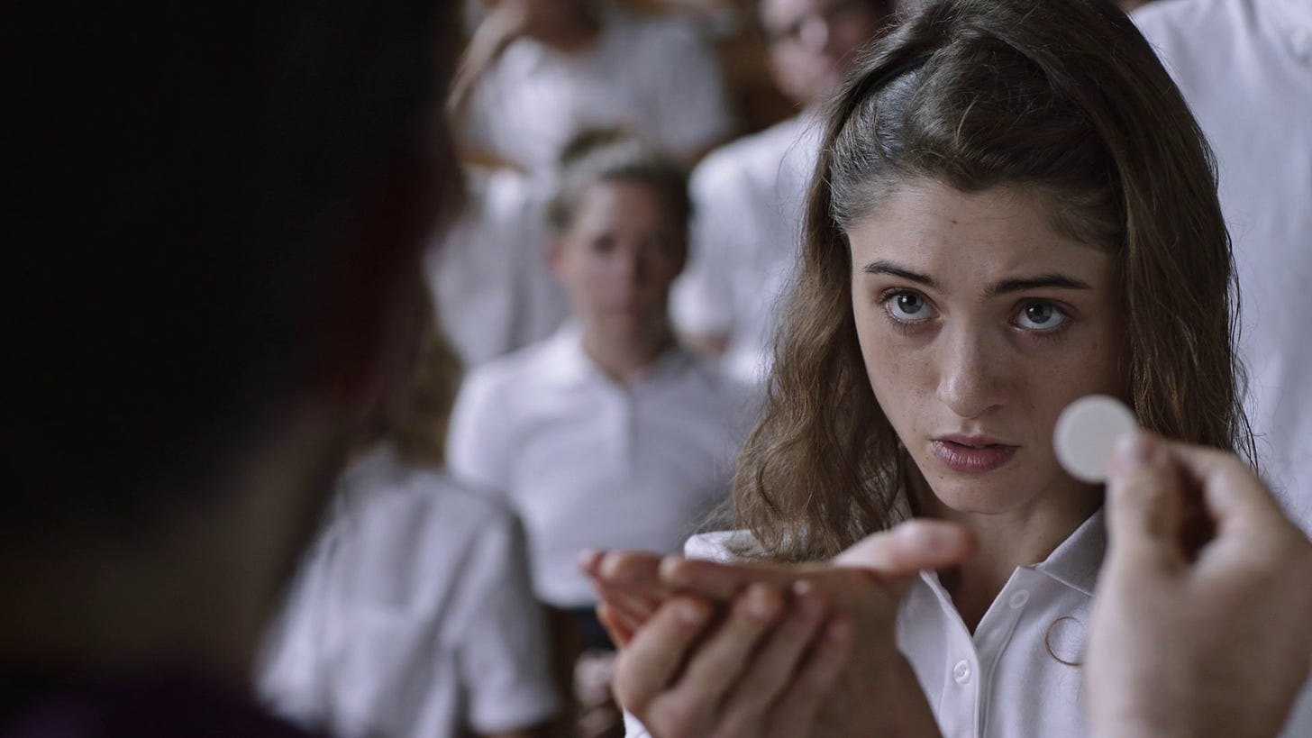 A white teenage girl, a character played by actress Natalia Dyer, holds out her hand among a group of uniformed Catholics behind her, waiting to receive the wafer of the Eucharist. It's a screenshot from the movie Yes, God, Yes