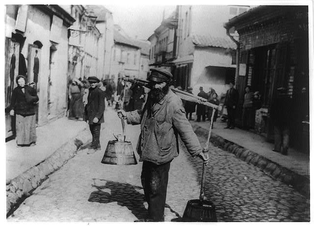 Black and white photo of a bearded man carrying a yoke with buckets of water on a crowded street.
