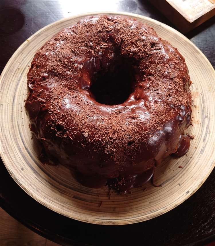 A giant glossy bundt cake (made in a ring mould) covered in chocolate ganache. and grated chocolate. It's on a circular bamboo plate against a dark wood background. The cake has dates underneath the ganache, giving it a rustic, bumpy look.