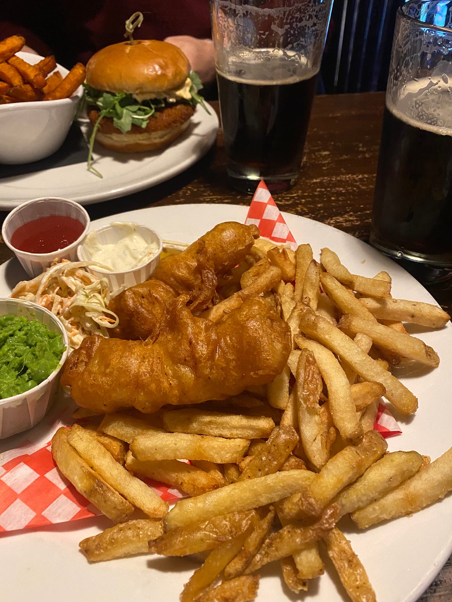 Two plates on a weathered wooden pub table, with glasses of dark beer beside them. The plate in the foreground has fish and chips with tartar sauce and ketchup in paper cups, next to a small pile of coleslaw and a slightly larger paper cup of mushy peas. The plate in the background has a veggie burger and a bowl of yam fries.