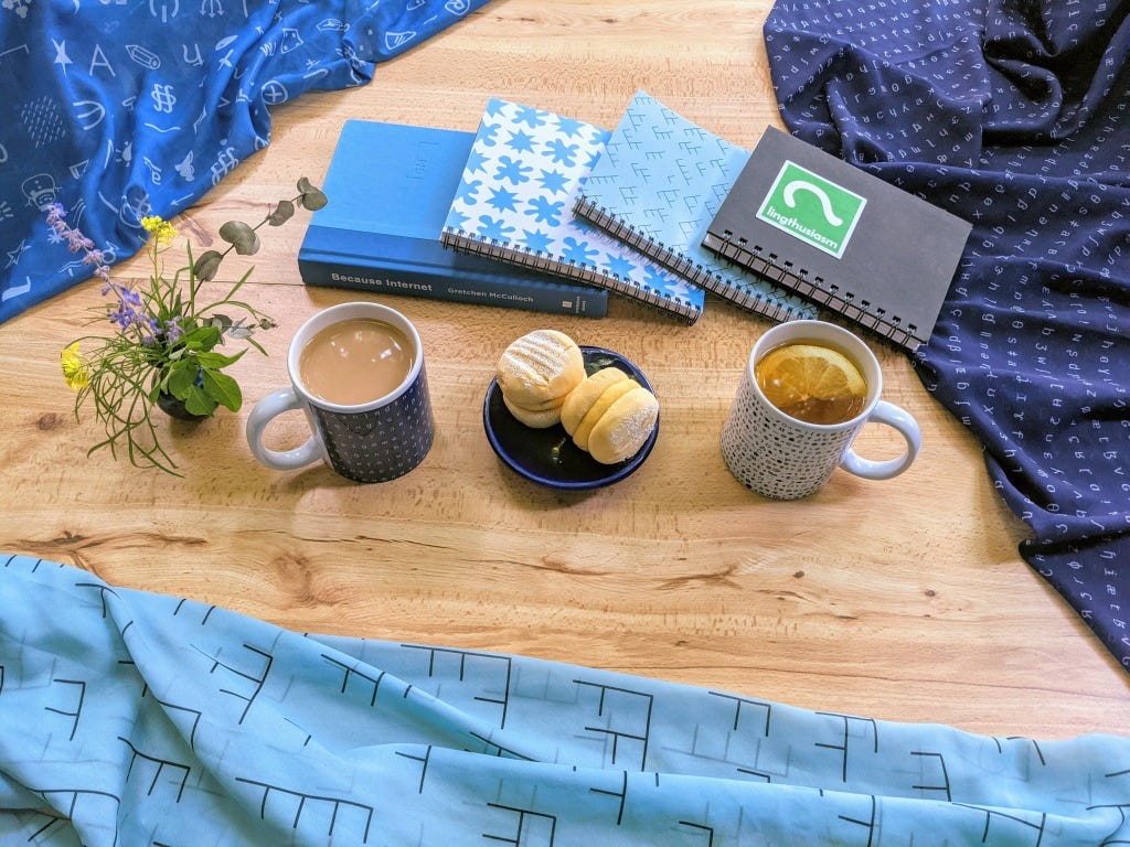 Lingthusiasm merch: Mugs of tea, pastries, Lingthusiasm notebooks, and BECAUSE INTERNET surrounded by blue Lingthusiasm tree diagram and esoteric symbols scarves. 