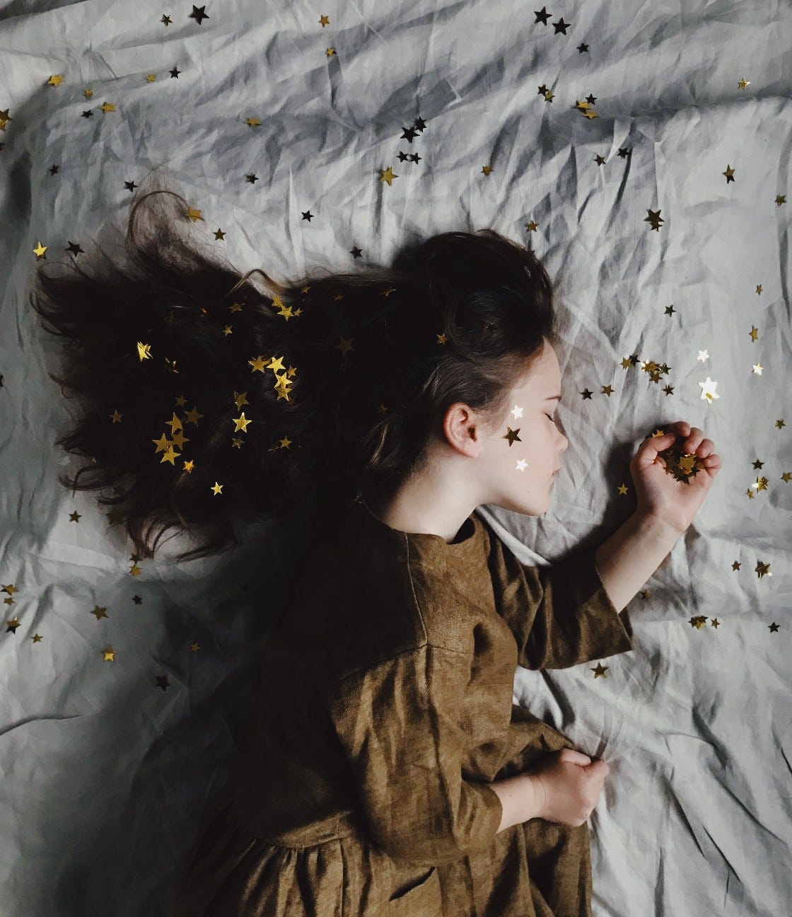 a young girl with long brown hair in a tan dress sleeps on a wrinkled muslin sheet. Small gold stars are sprinkled through her hair and on the sheet, and she holds some in one hand.