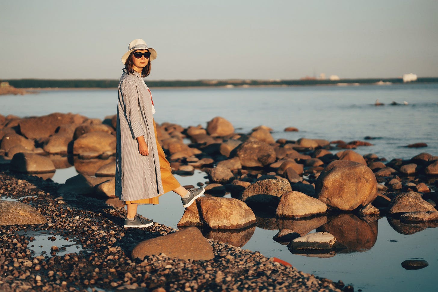 A woman in sunglasses and a long coat stands casually beside a river.