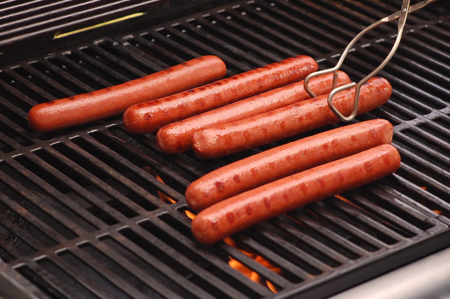 6 long hot dogs on a clean grill, one being turned with some metal tongs