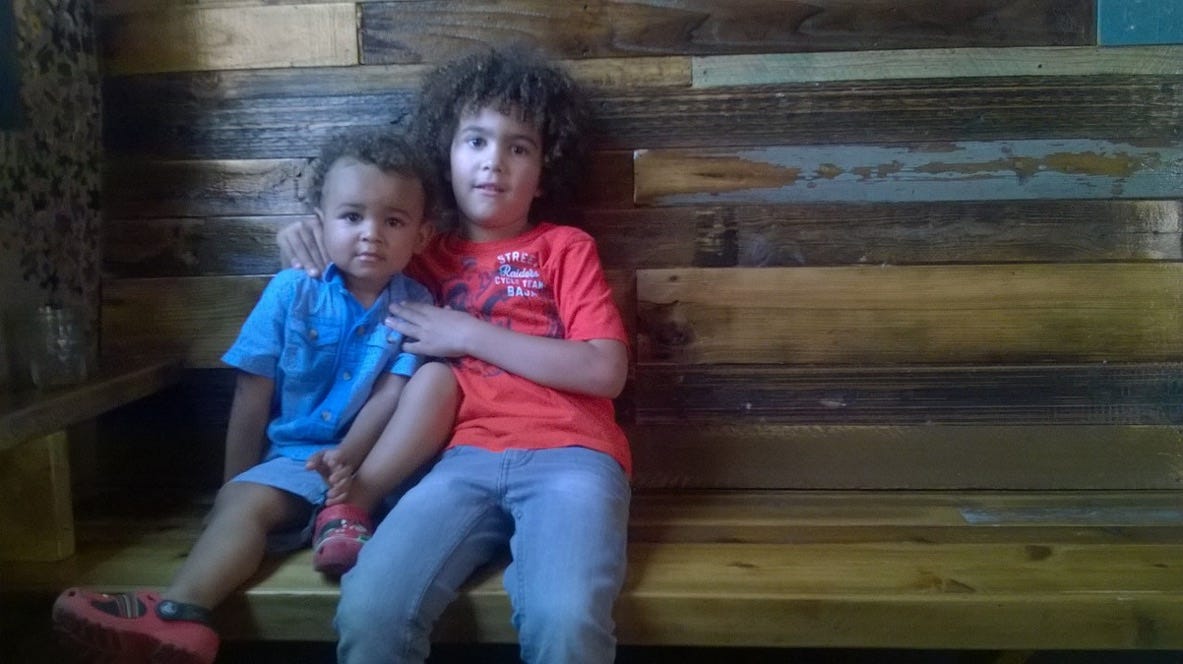 Two gorgeous brown curly-haired boys sitting on a wooden bench, with reclaimed wood behind them.