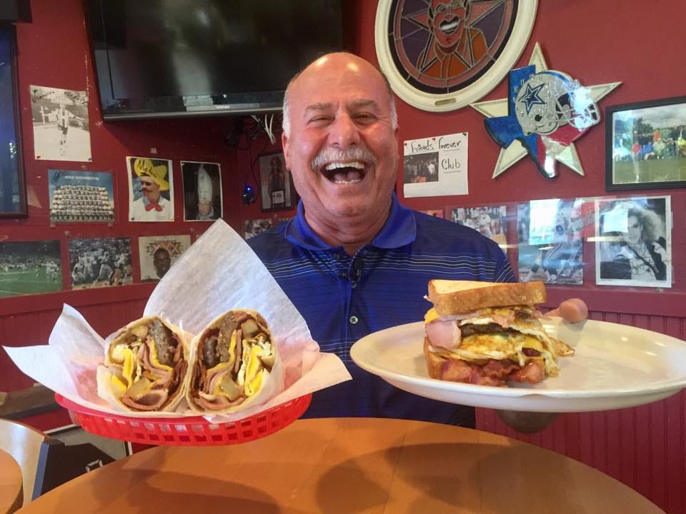 Coppell Deli owner Jay Khorrami smiles broadly while displaying two obscenely large sandwiches.