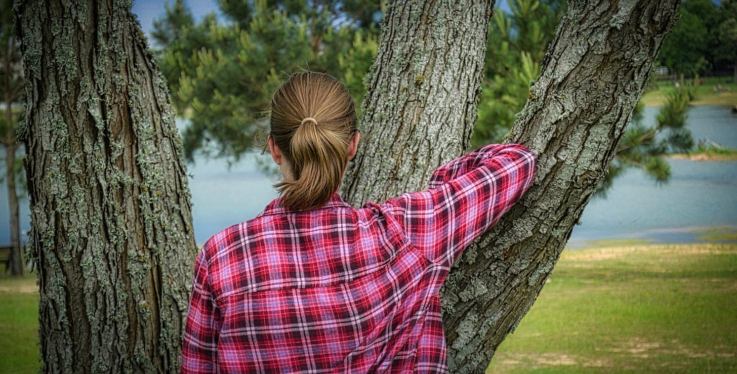 Young woman with a pony tail facing the lake in a red flannel shirt and leaning against a tree with blue sky and bits of lake in the background