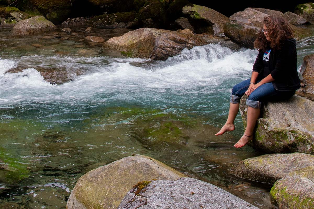 woman with wavy brown hair sits on a boulder by the river's edge, bare feet dangling over the clear water, looking down at the water as the cold blue current rushes past in a stream of whitewater