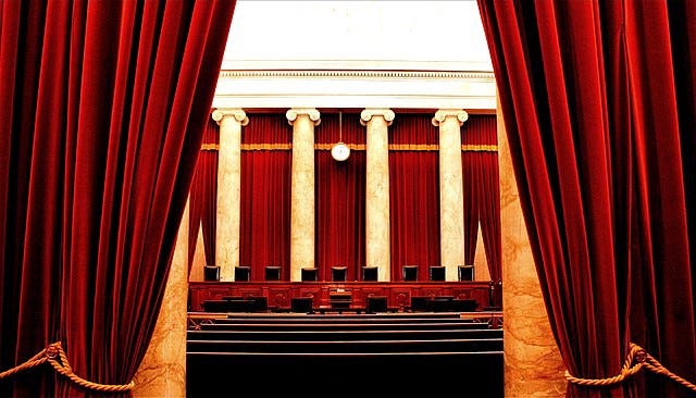 Image description: Inside of the Supreme Court of the United States, showing red velvet curtains on both the left and right sides of the image, drawn back to reveal nine dark brown seats behind a wooden podium. They are backed by four marble pillars and more red velvet curtains. Before the nine seats are more dark seats situated on a lower level, with dark pews behind them.