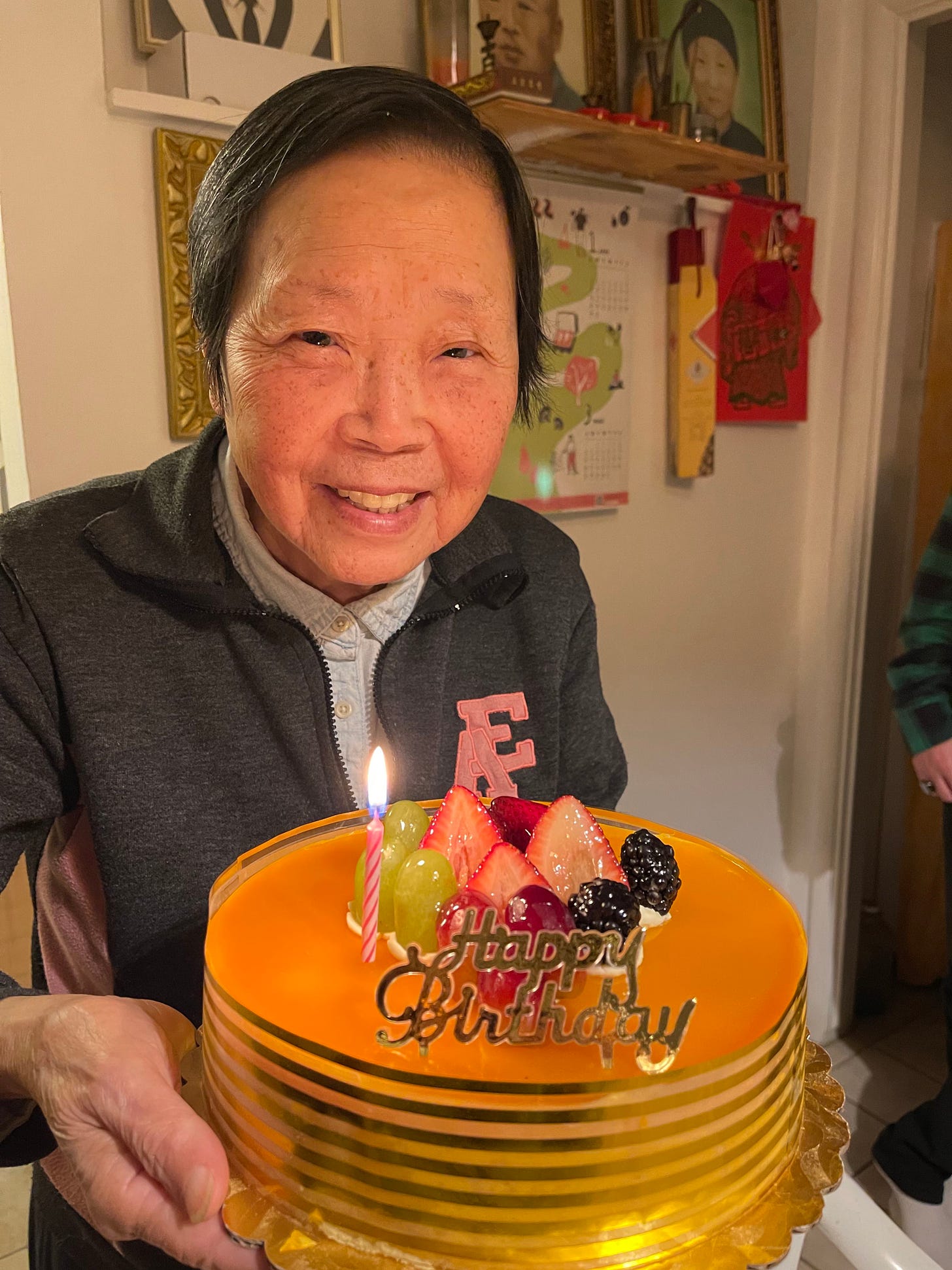Photo of Mama Yang smiling to the camera and holding a big orange cake with the words "Happy Birthday" on top