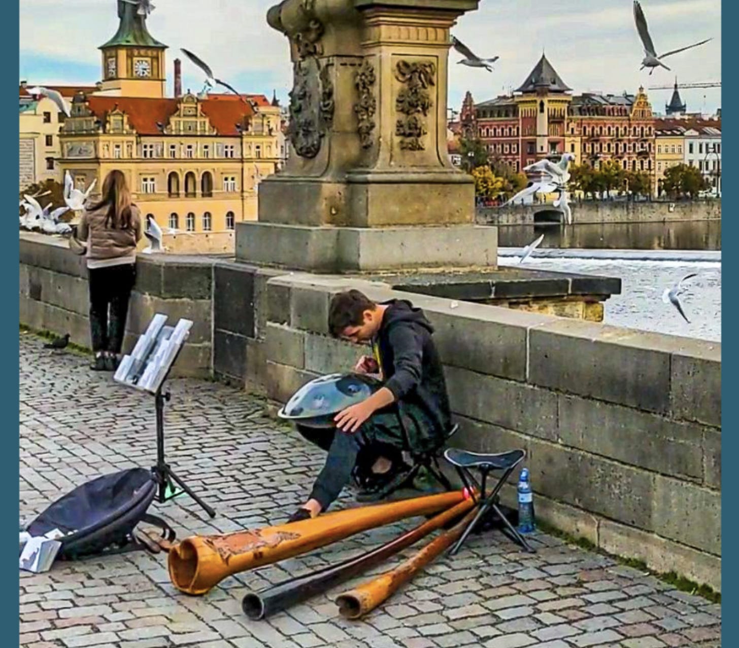 A young man plays a metal drum and enormous wooden horns on the Charles Bridge as seagulls circle overhead.