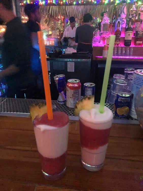 two frozen alcoholic beverages sit on a wooden bar table, parts red and yellow, with one green straw, and one orange straw.
