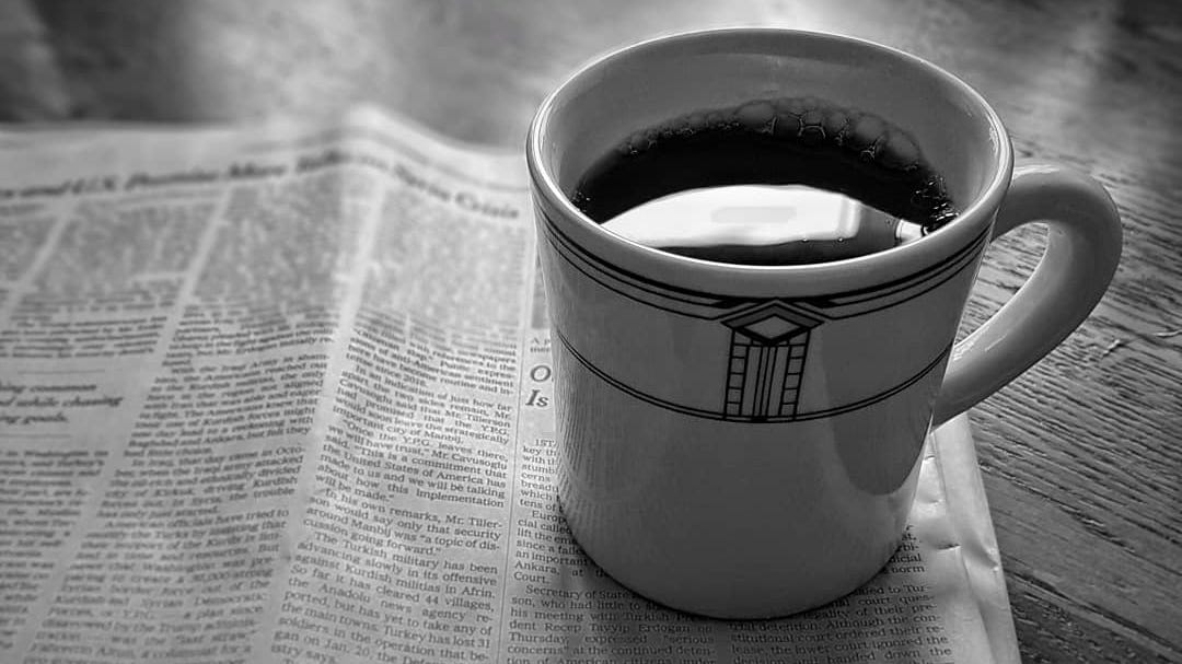 A diner mug full of coffee rests on a newspaper atop a wooden table.