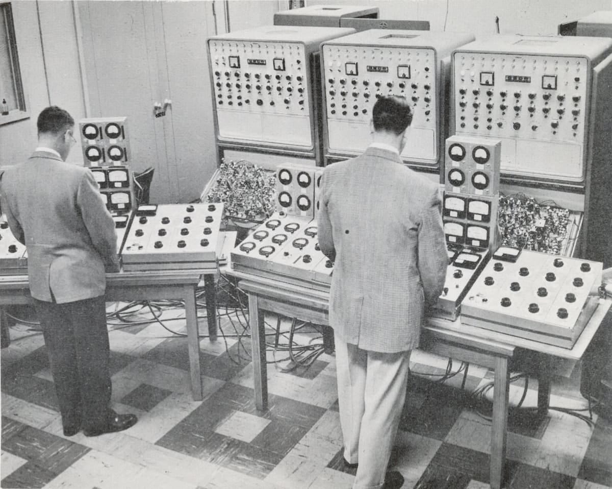 Black and white photo of two men in suits standing in front of complex control panels, with a 1950s computer behind.