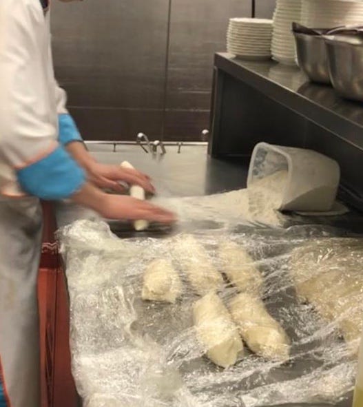 A chef in a professional kitchen quickly rolls rolls out noodle dough on a stainless steel surface