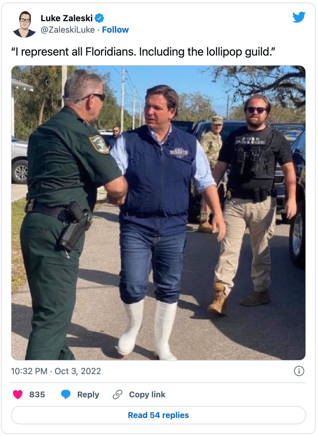 Tweet by Luke Zaleski that reads: "I represent all Floridians. Including the lollipop guild.” with a picture of Ron DeSantis shaking hands with a Florida cop of some kind, while dressed like Violet Beauregarde (post-blueberry gum) and wearing hilarious shiny white knee-high rubber shrimp boots like the real good ol’ boy that he manifestly isn’t.