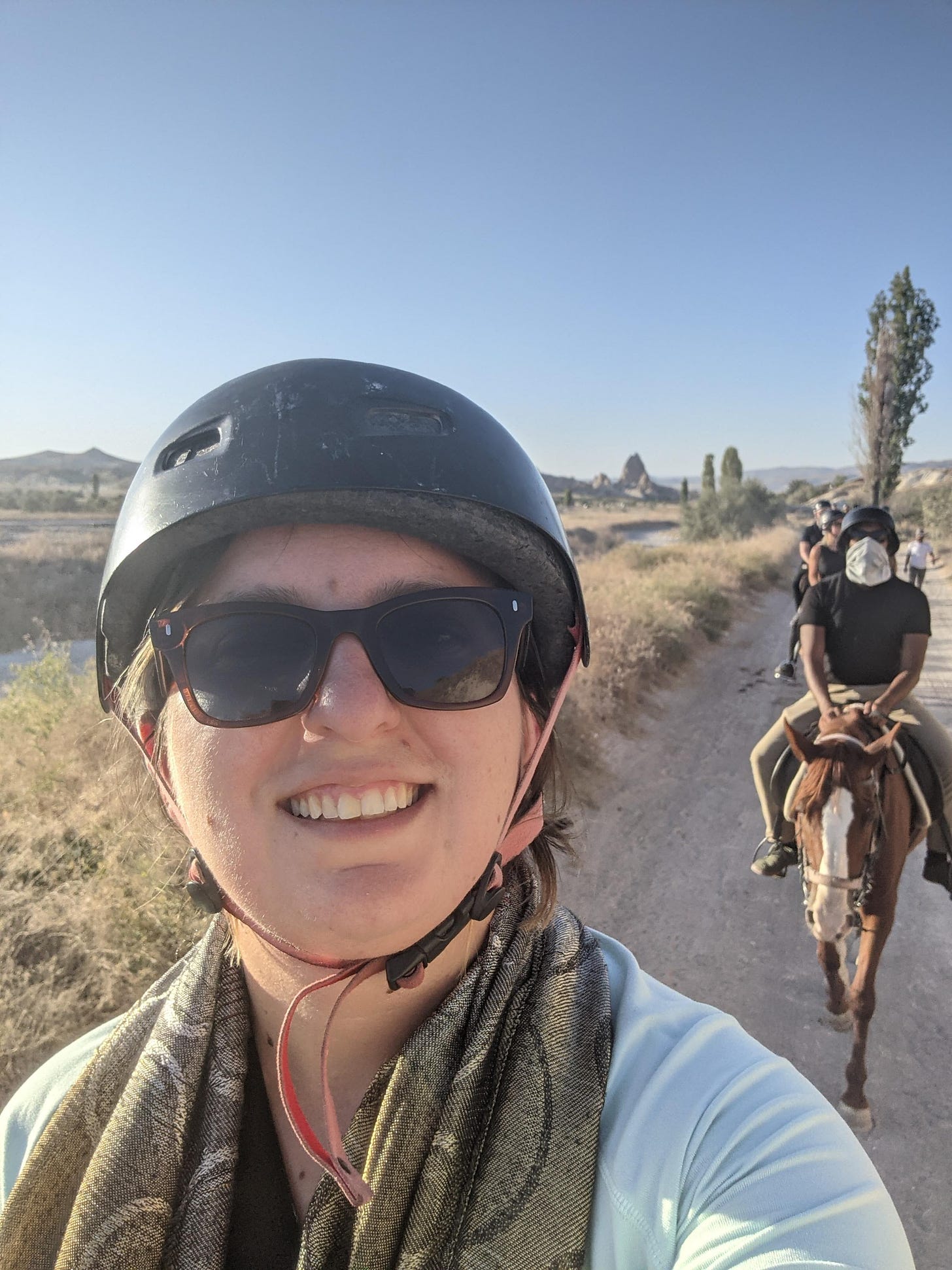 Selfie on horseback, wearing helmet, with a column of horses behind me, including Aseef riding a horse, and the Cappadocia landscape
