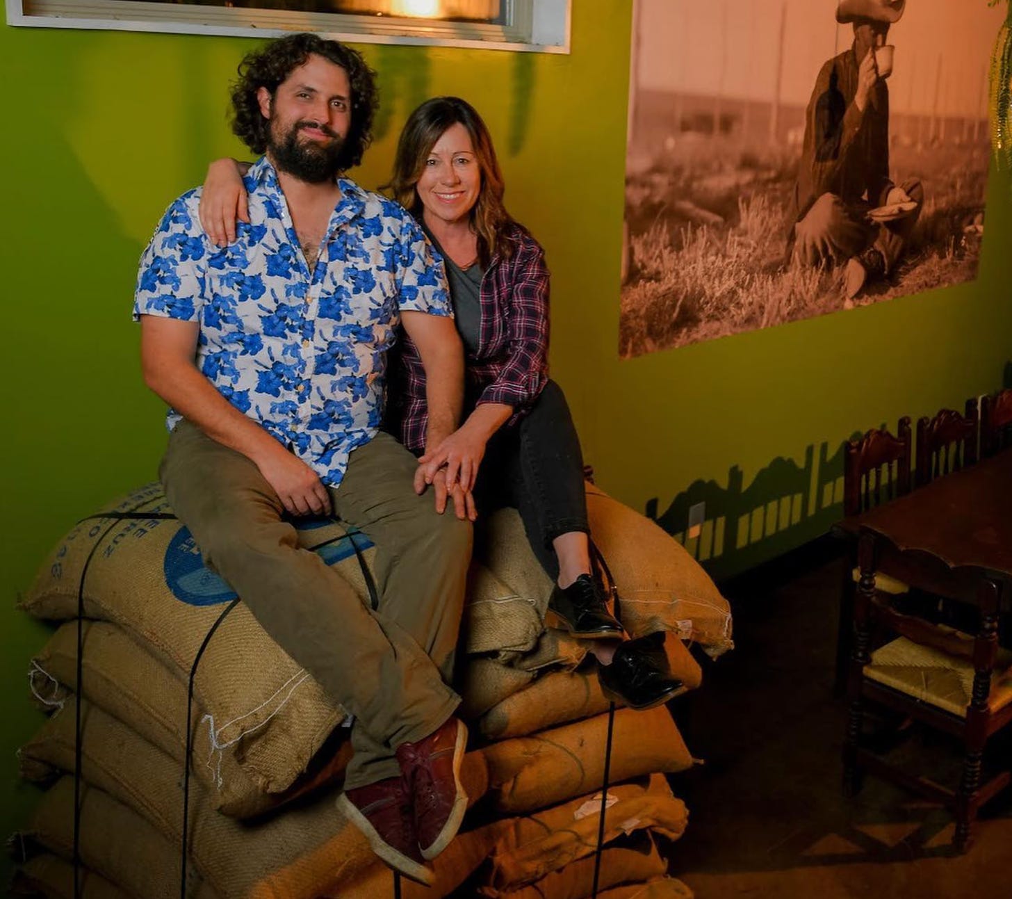 Samantha and Weston Nawrocki, owners of Manzanita Roasting, sitting on a stack of coffee bags in front of a green wall.