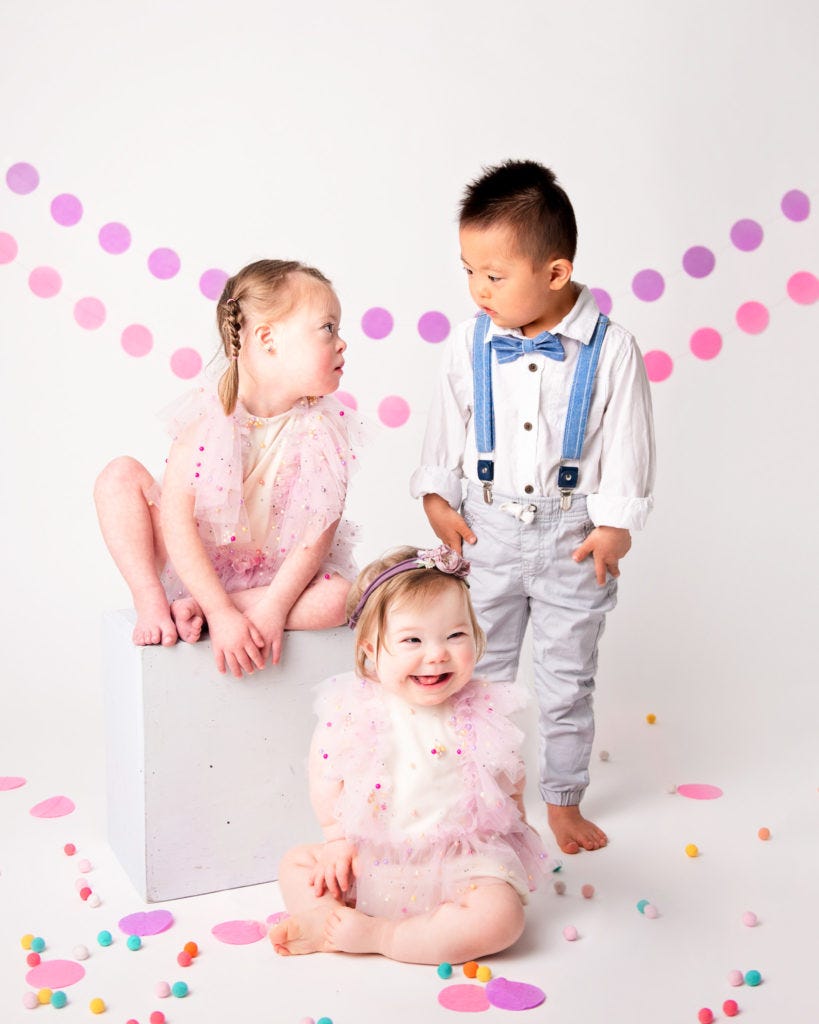 Three children with Downs Syndrome smiling in white studio wih colorful decorations.