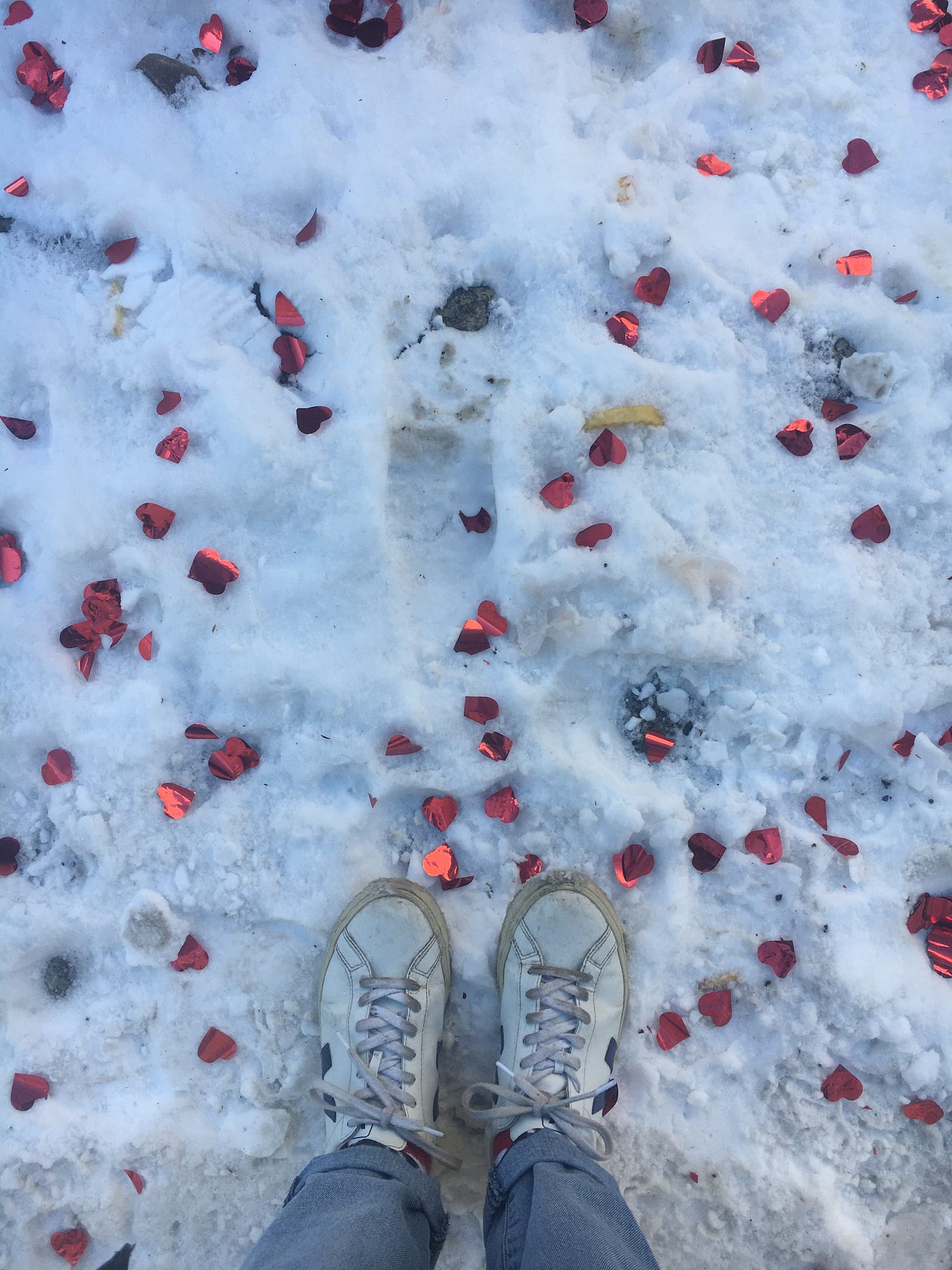 Schneebedeckter Boden, auf dem Herzkonfetti liegt. Unten ein paar Beine in heller Jeans und dreckigen weißen Turnschuhen.