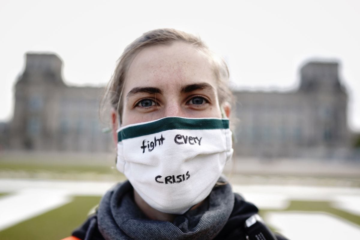 A climate activist stands in front of an official building. She is wearing a facemask that says 'fight every crisis' and looking straight at the camera.