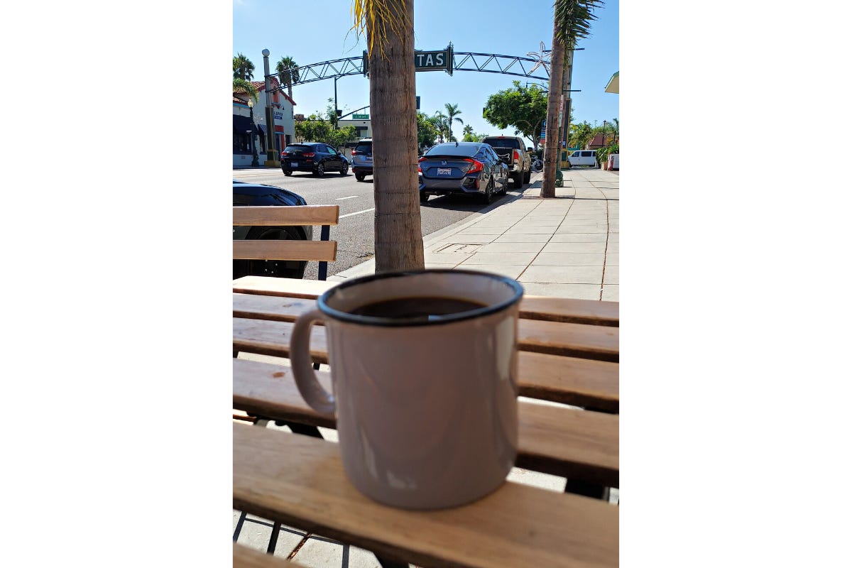 A blurry coffee mug filled with coffee sits on a patio table in the foreground. Rising up right behind it is the trunk of a palm tree which splits and obstructs the view of the city's historical arching sign over the road. You can just see the white -TAS letters from the name Encinitas to the right of the trunk.