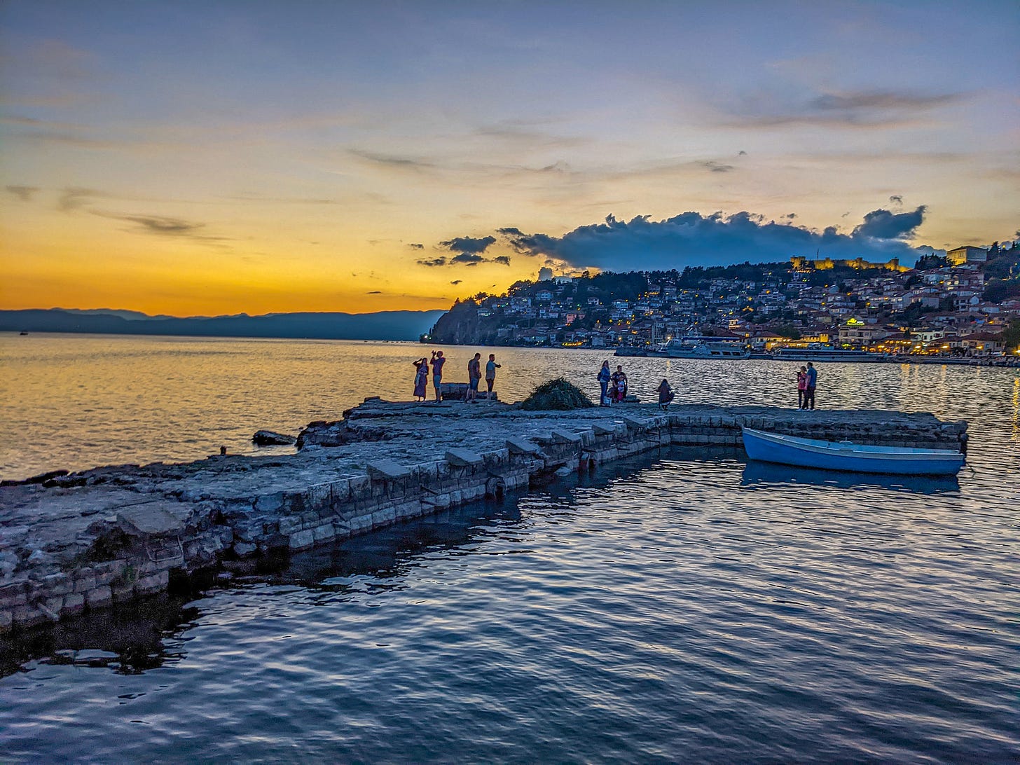 The town of Ohrid from the pier during sunset. The lights of the town are lit up and people on the pier are silhouetted.