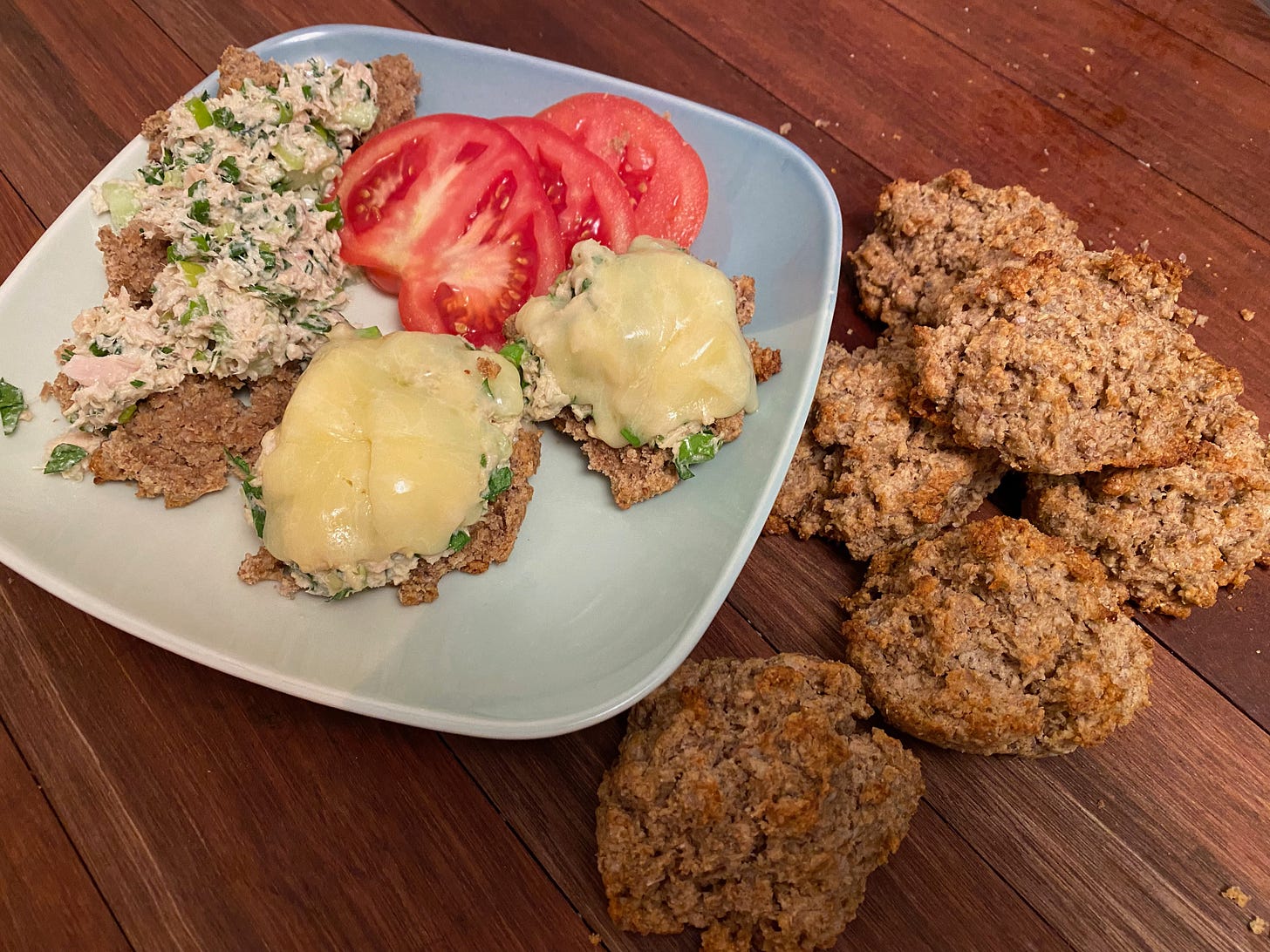 A square plate holds a pile of tuna salad, two halves of a rye biscuit made into a tuna melt, and several tomato slices. A pile of golden brown rye biscuits sits on the wooden counter beside the plate.