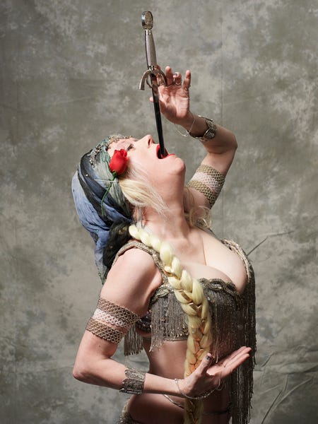 Foto de uma mulher loira de meia-idade vestida com roupa de dança do ventre, uma rosa presa na altura da orelha, engolindo uma espada