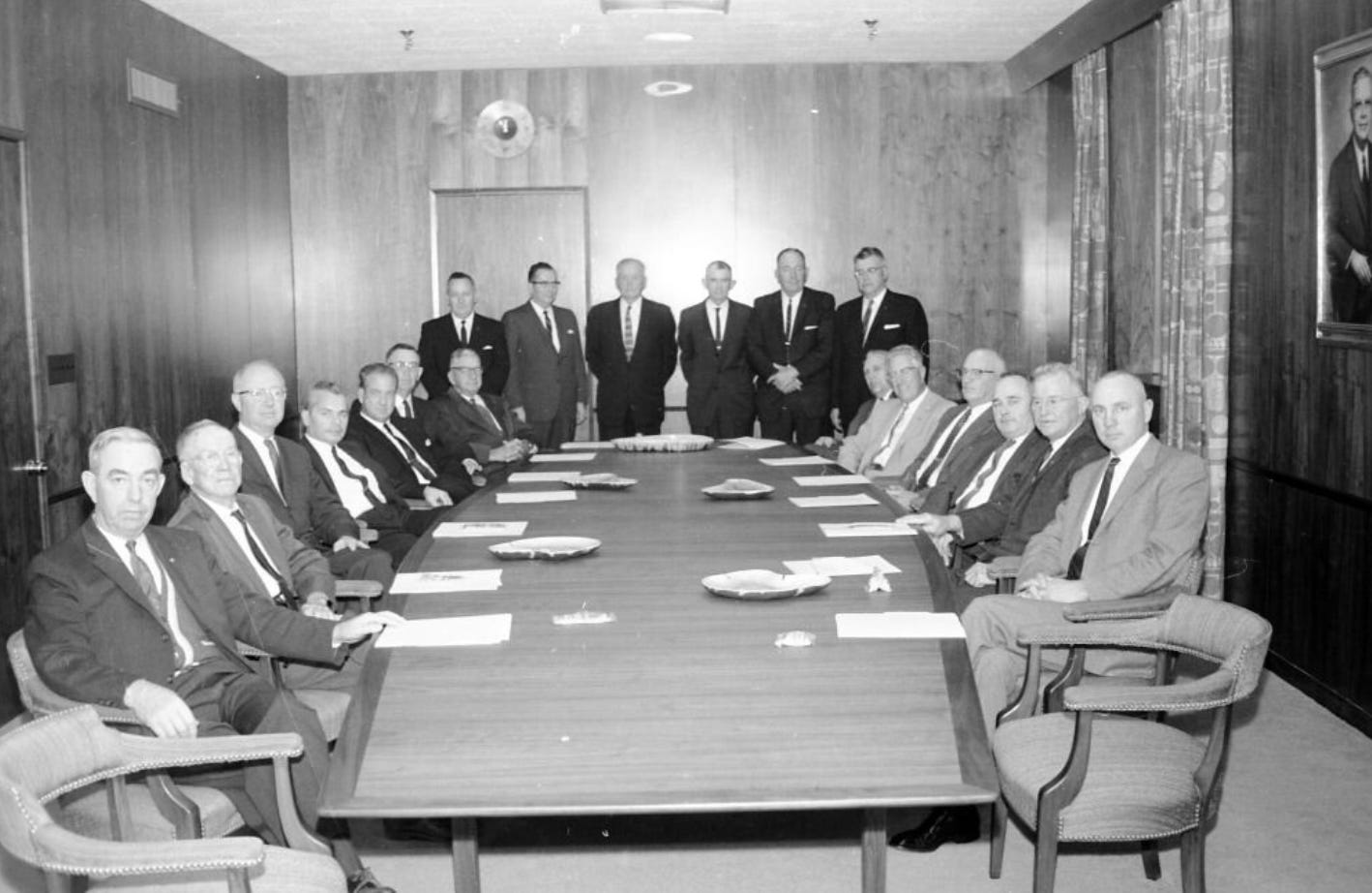 Another picture of men in suits, this one they're all fanned out around a very large table in a wood paneled room