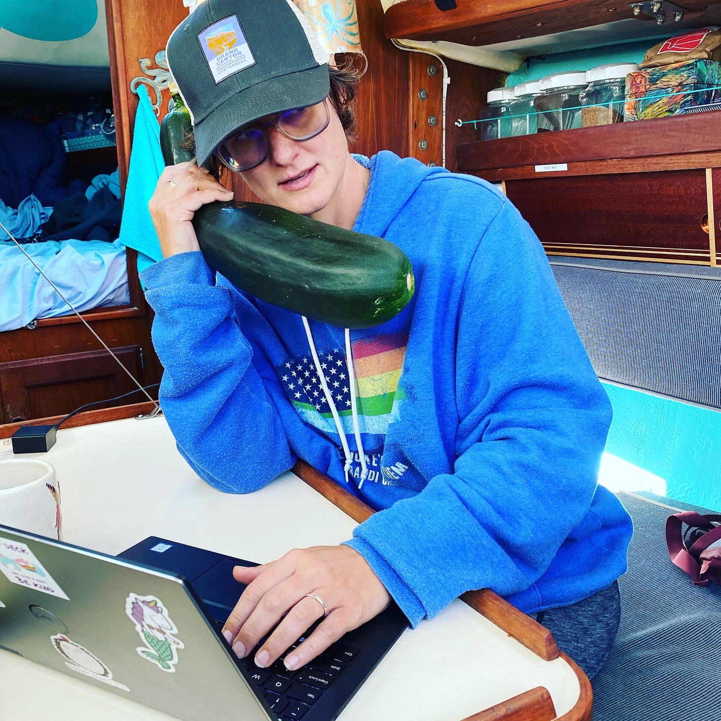 Skipper Jenn sitting at a table in her boat holding a giant zucchini to her ear like she is on the phone.
