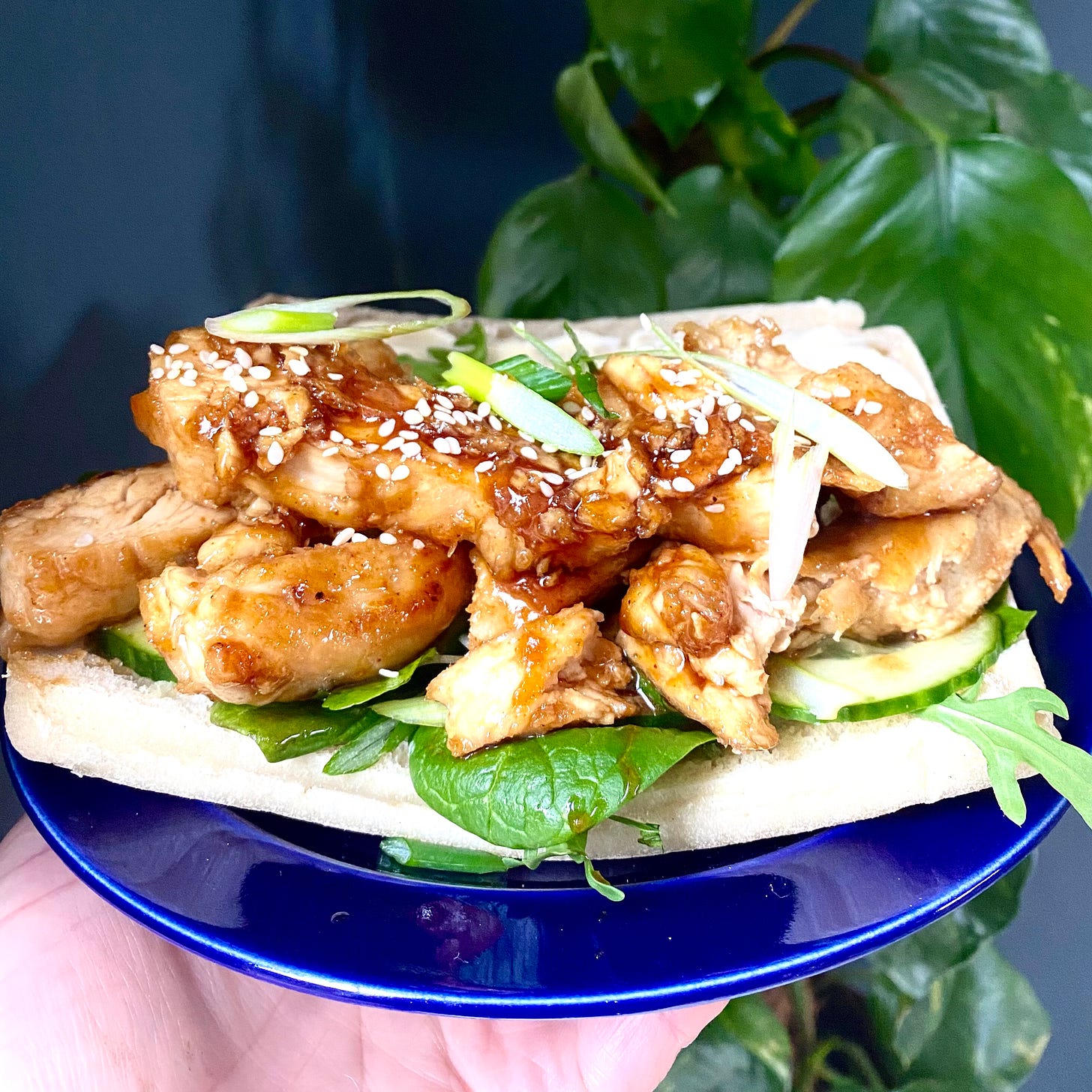 Blue plate with a sandwich on top, filled with Korean fried chicken, shredded spring onions, spinach and cucumber