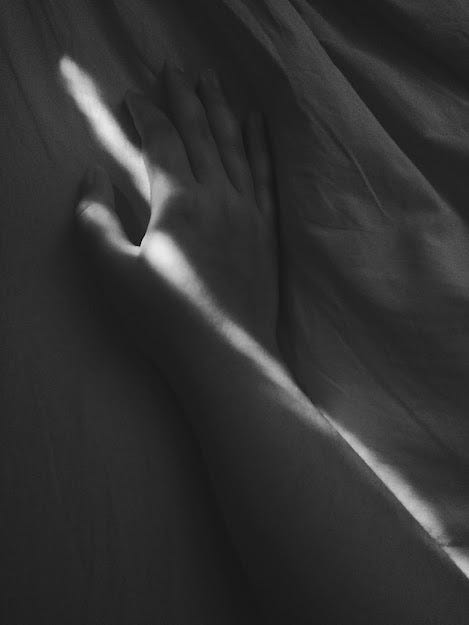 Imagem em preto e branco de uma mão sobre o lençol da cama. Uma fresta de luz cruza a mão.