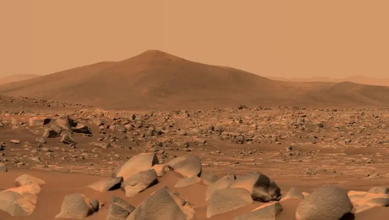 A rust red Martian landscape, rocks in the foreground, an low, flat hill in the background.