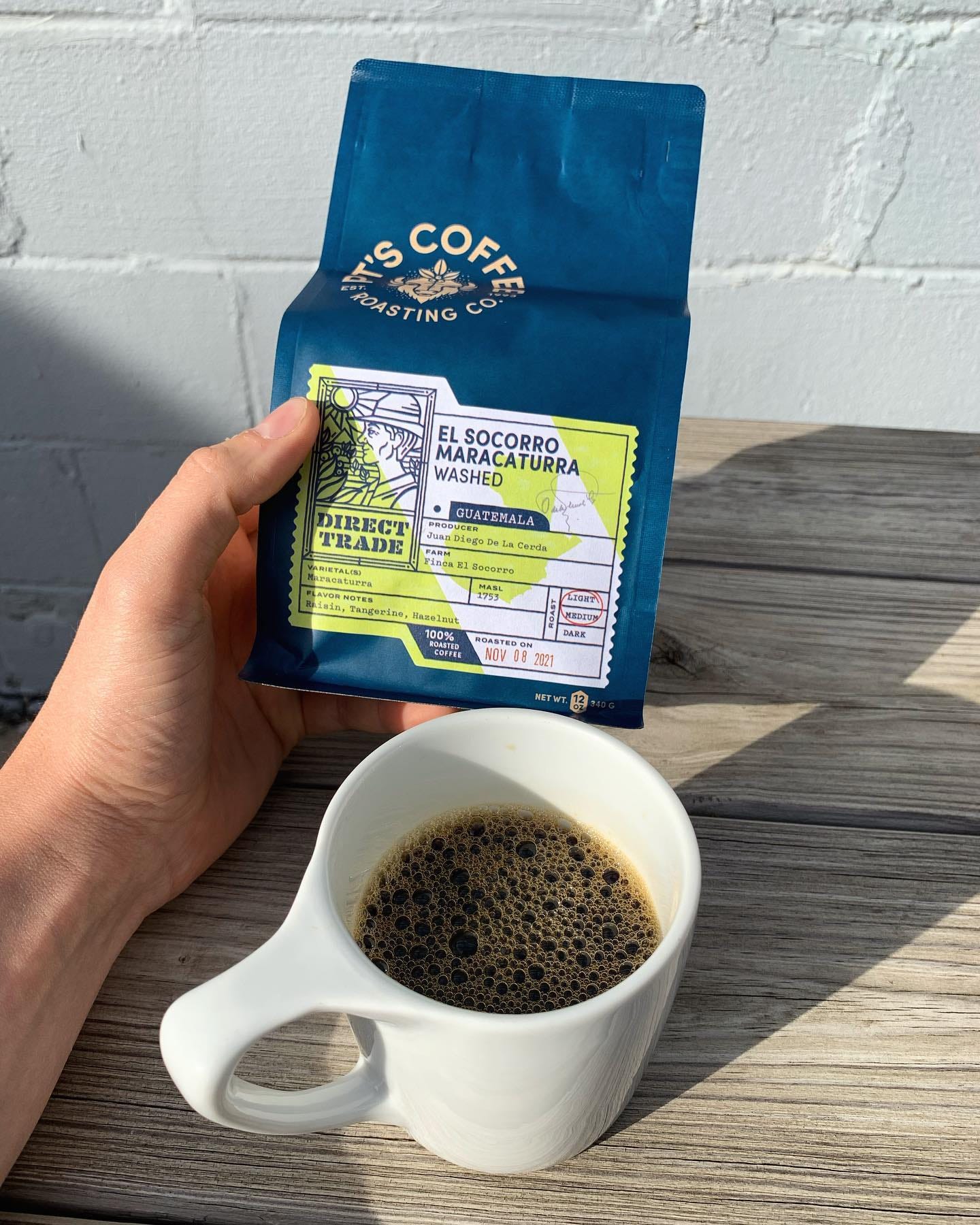 A hand holding a blue bag of PT's Coffee roasting company washed Guatemala coffee beans in front of a white concrete wall. A full coffee cup sits on a picnic table below.