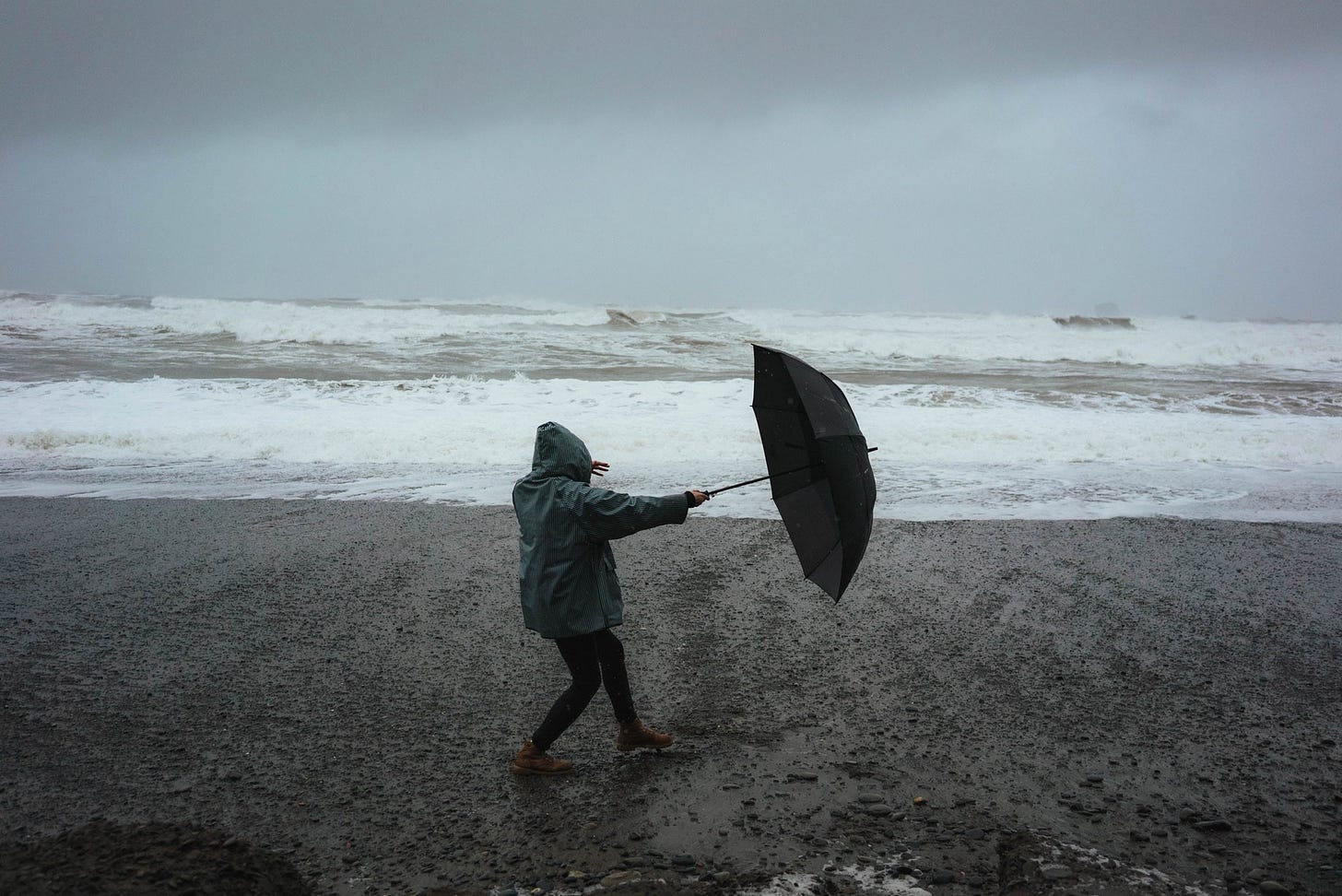Person in dark green rain coat on a beach near the water. It's a dark, stormy situation, and the person is holding an umbrella which they are trying to hold against the wind, which is pulling them to the right.