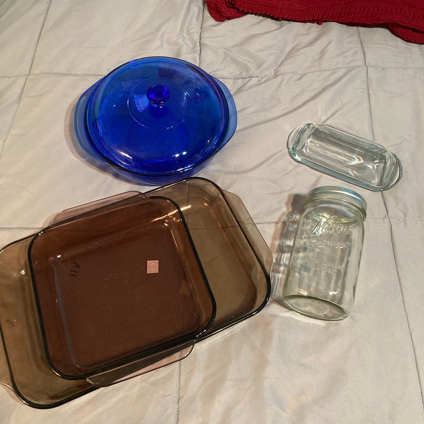 Image Description: A round cobalt blue baking dish with a lid, a clear glass butter dish, a clear glass wide mouth Kerr mason jar, a rectangular amber glass baking dish nested with a square purple glass baking dish all lay on a gray quilted bedspread. 
