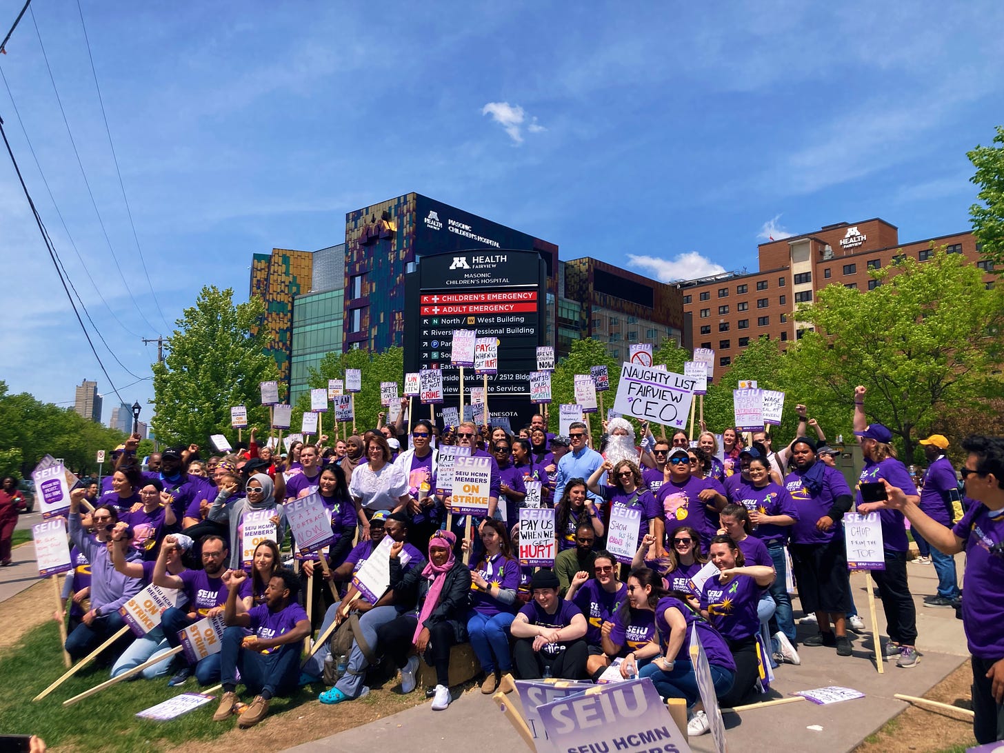 a large crowd of people wearing purple tee shirts and holding picket signs gather in front of the m health fairview riverside hospital sign on the sidewalk