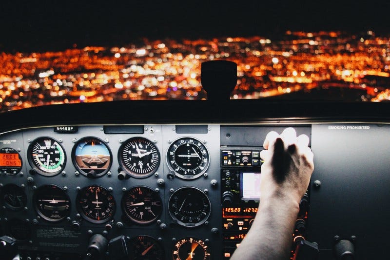 A pilot grabbing the controller of the plane over a panel with flight instruments and a city night landscape outside