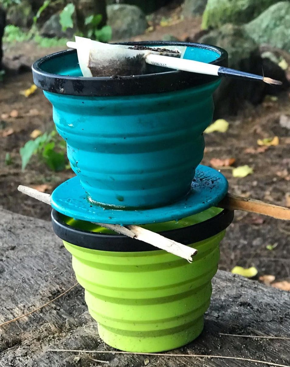 A collapsible teal coffee pour over propped over a green collapsible cup with random sticks. A coffee filter filled with coffee sits in the top. Foliage and trail are blurred in the background.
